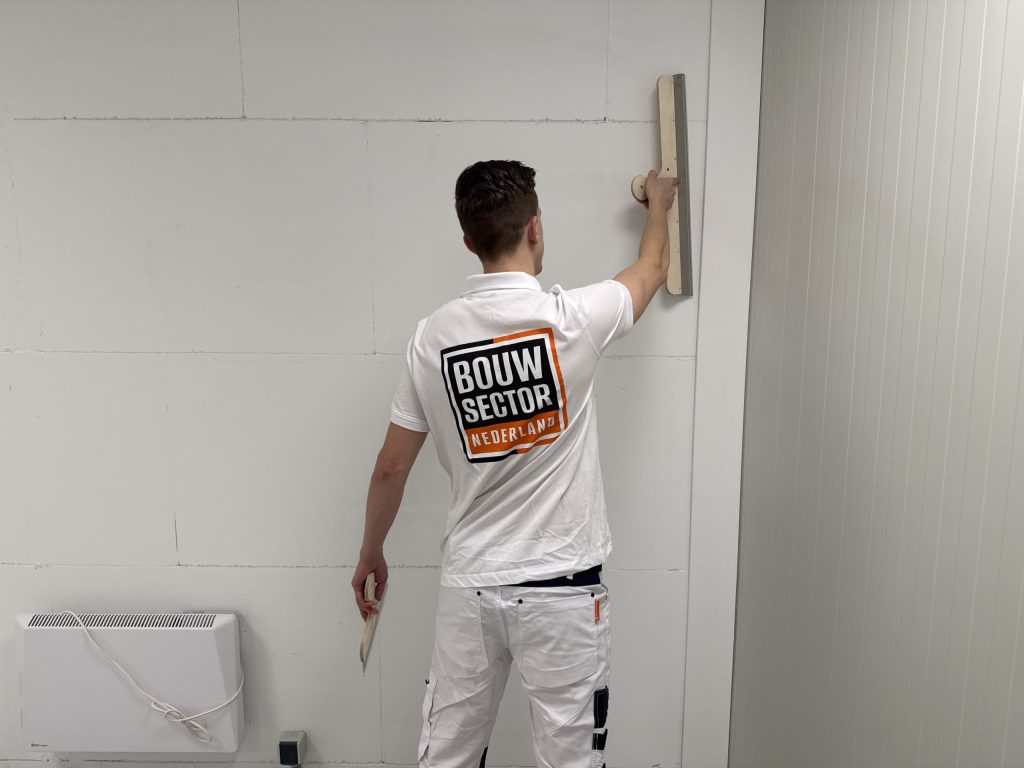 Stukadoor van Bouwsector Nederland in wit werktenue zet een lange spaan verticaal tegen de bovenkant van een wand