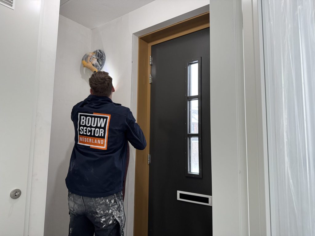 Vakman van Bouwsector Nederland schuurt met een wandschuurmachine de muur boven een voordeur met zijlicht